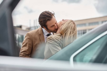 elegant businessman in formal wear embracing and kissing blonde woman near car on urban street