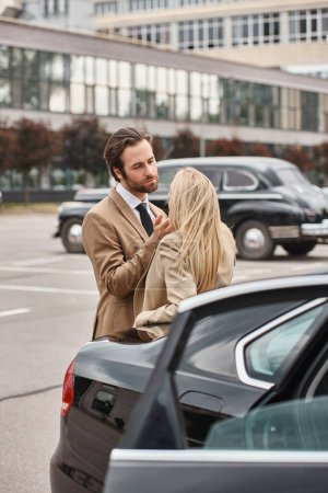 handsome businessman in elegant formal wear looking at blonde woman near car on urban street