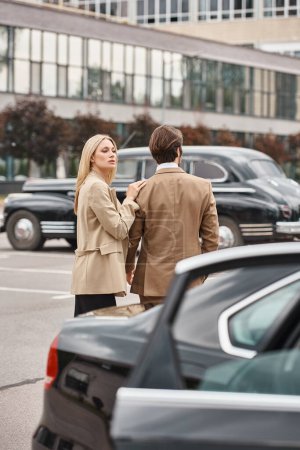 attractive blonde businesswoman in formal wear touching shoulder of elegant man near car on street
