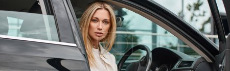 femme blonde élégante et expressive assise dans une voiture moderne et regardant la caméra, bannière horizontale