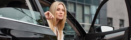 mujer rubia moderna en ropa casual sentado en el coche y mirando hacia otro lado en la calle urbana, pancarta