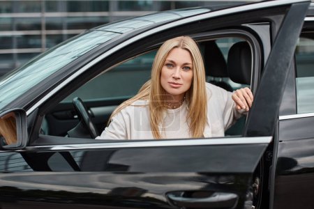 blonde woman in trendy casual clothes sitting in modern car and looking at camera on city street