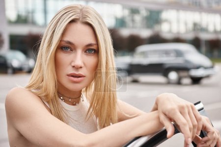Téléchargez les photos : Attrayant et confiant femme blonde regardant caméra tout en se tenant près de la voiture sur la rue de la ville - en image libre de droit