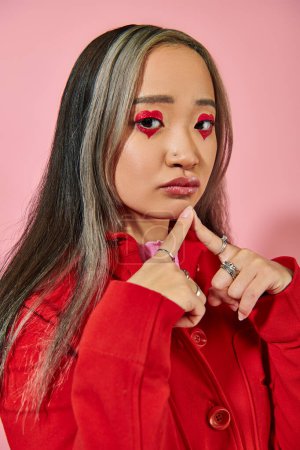 pensive young asian woman with heart shaped eye makeup and dyed hair looking at camera on pink
