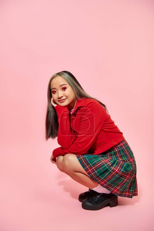 full length of happy asian woman with heart shaped eye makeup and plaid skirt sitting on pink