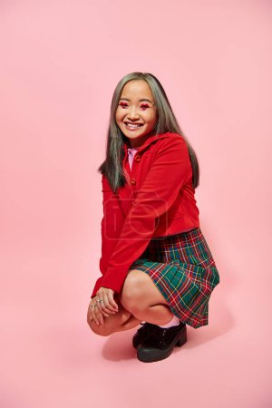 full length of cheerful asian woman with heart shaped eye makeup and plaid skirt sitting on pink