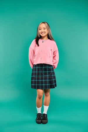full length of cheerful asian woman in pink sweatshirt and plaid skirt standing on blue backdrop