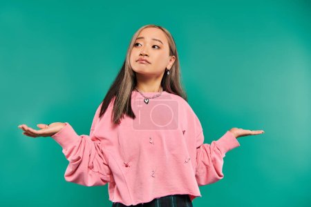 portrait of confused asian woman in pink sweatshirt and heart shaped necklace on blue backdrop