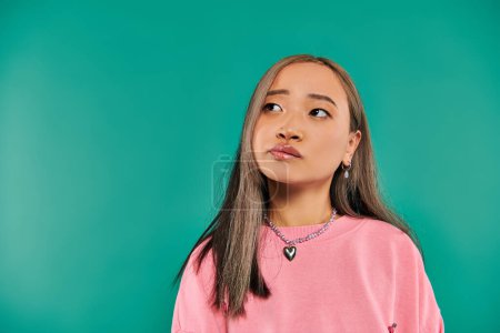 portrait of pensive asian woman in pink sweatshirt and heart shaped necklace on blue backdrop