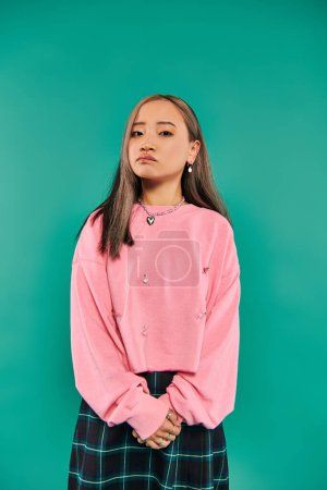 portrait of pensive asian woman in pink sweatshirt and plaid skirt standing on blue backdrop