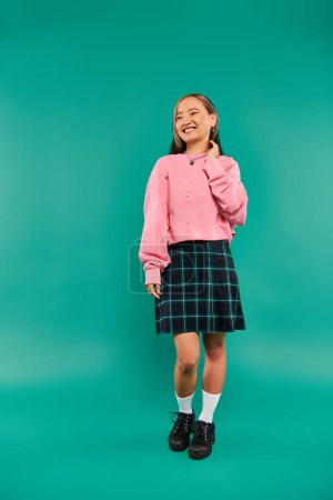 full length of happy young asian woman in pink sweatshirt and plaid skirt standing on pink backdrop