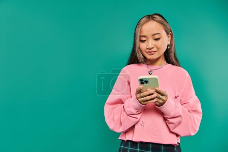 portrait of cheerful asian woman in pink sweatshirt using smartphone on turquoise backdrop