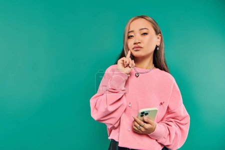 portrait of pensive young asian woman in pink sweatshirt using smartphone on turquoise backdrop