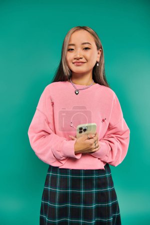 portrait of happy young asian woman in pink sweatshirt and plaid skirt using smartphone on turquoise