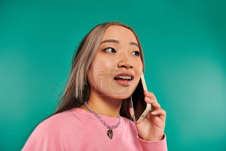 Porträt einer hübschen jungen Asiatin im rosafarbenen Sweatshirt, die auf einem Smartphone vor türkisfarbenem Hintergrund spricht