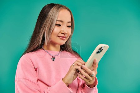 portrait of pleased young asian woman in pink sweatshirt and plaid skirt using smartphone on blue
