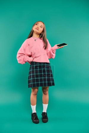 attractive young asian woman in sweatshirt and plaid skirt holding smartphone on turquoise backdrop