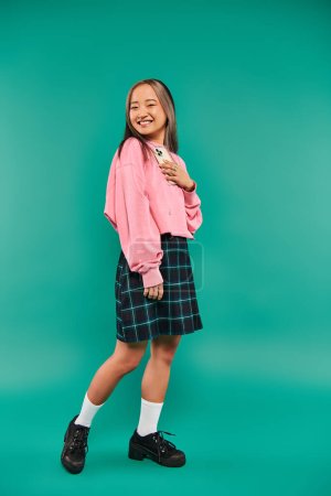 happy young asian woman in sweatshirt and plaid skirt holding smartphone on turquoise backdrop