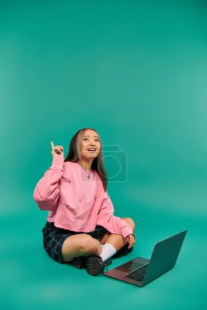 Foto de Feliz mujer asiática en sudadera y falda a cuadros teniendo idea mientras se utiliza el ordenador portátil en el telón de fondo turquesa - Imagen libre de derechos