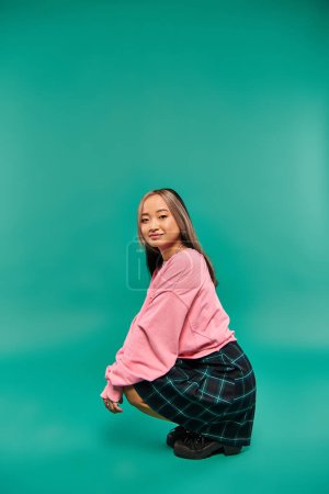longitud completa de feliz joven asiático mujer en rosa sudadera y falda a cuadros sentado en rosa telón de fondo