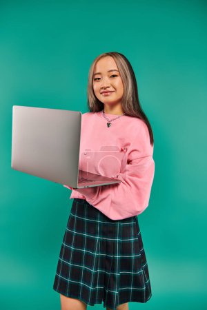 Foto de Feliz asiático chica en rosa sudadera usando portátil mientras de pie en turquesa telón de fondo, la creación de redes - Imagen libre de derechos