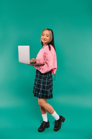 Foto de Complacido chica asiática en sudadera rosa usando el ordenador portátil, mientras que de pie en el telón de fondo turquesa, la creación de redes - Imagen libre de derechos