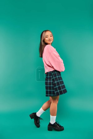 full length of pleased asian girl in pink sweatshirt and plaid skirt standing on pink backdrop