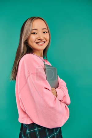 Foto de Alegre asiático chica en rosa sudadera holding laptop mientras de pie en turquesa telón de fondo - Imagen libre de derechos