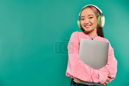 joyful asian girl in wireless headphones holding laptop while standing on turquoise backdrop