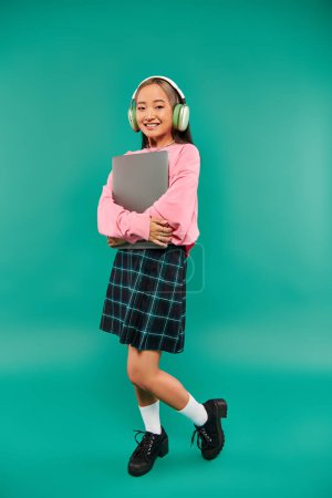 joyful asian girl in wireless headphones holding laptop and standing on turquoise backdrop