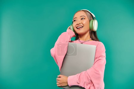 positive asian girl in wireless headphones holding laptop while standing on turquoise backdrop