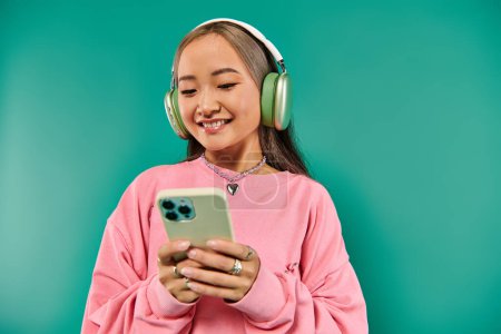 lustige asiatische Mädchen in drahtlosen Kopfhörern Musik hören und mit Smartphone auf türkisfarbenem Hintergrund