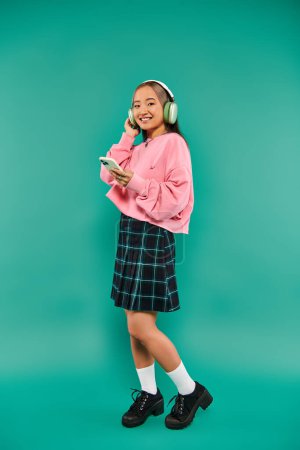 Foto de Alegre asiático chica en inalámbrico auriculares escuchar música y el uso de teléfono inteligente en turquesa - Imagen libre de derechos