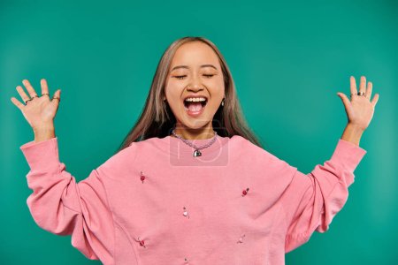 portrait of excited and young asian girl in pink sweatshirt posing on turquoise background