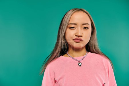 portrait of skeptical and young asian girl in pink sweatshirt posing on turquoise background