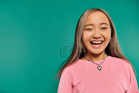 portrait of cheerful and young asian girl in pink sweatshirt posing on turquoise background
