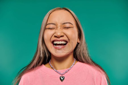 portrait of positive and young asian girl in pink sweatshirt posing on turquoise background