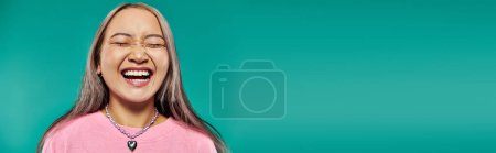 portrait of cheerful and young asian girl in pink sweatshirt posing on turquoise background, banner