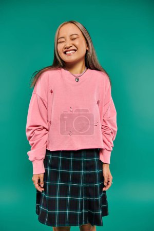 Porträt eines zufriedenen und jungen asiatischen Mädchens in rosa Sweatshirt posiert auf türkisfarbenem Hintergrund