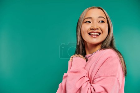Porträt eines fröhlichen und jungen asiatischen Mädchens in rosa Sweatshirt posiert auf türkisfarbenem Hintergrund