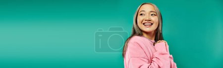 portrait of cheerful and young asian girl in pink sweatshirt posing on turquoise background, banner