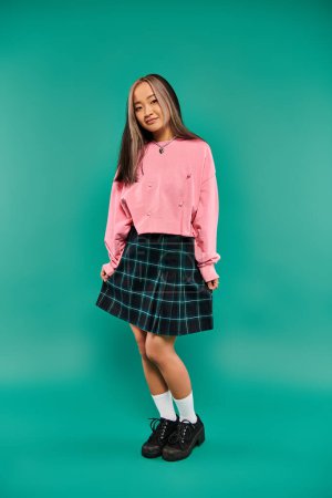 full length of cheerful and young asian girl in pink sweatshirt posing on turquoise background