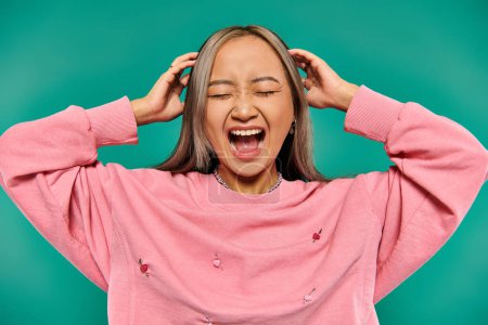 portrait of emotional young asian girl in pink sweatshirt screaming on turquoise background
