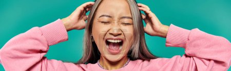 portrait of emotional young asian girl in pink sweatshirt screaming on turquoise background, banner