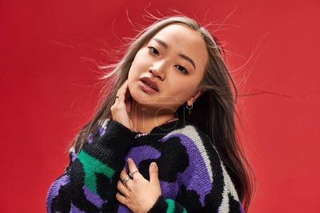 alluring young asian woman in vibrant sweater with animal print touching cheek on red backdrop