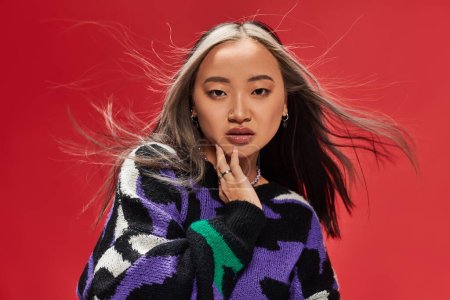 charming young asian woman in vibrant sweater with animal print touching cheek on red backdrop