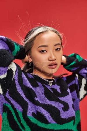 charming young asian woman in vibrant sweater with animal print adjusting hair on red backdrop
