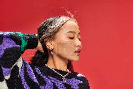 alluring young asian woman in vibrant sweater with animal print adjusting hair on red backdrop