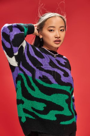attractive young asian woman in vibrant sweater with animal print adjusting hair on red backdrop
