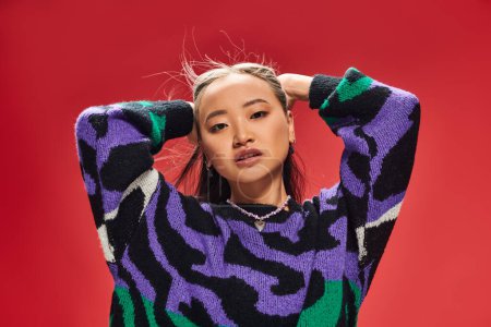 beautiful young asian woman in vibrant sweater with animal print adjusting hair on red backdrop
